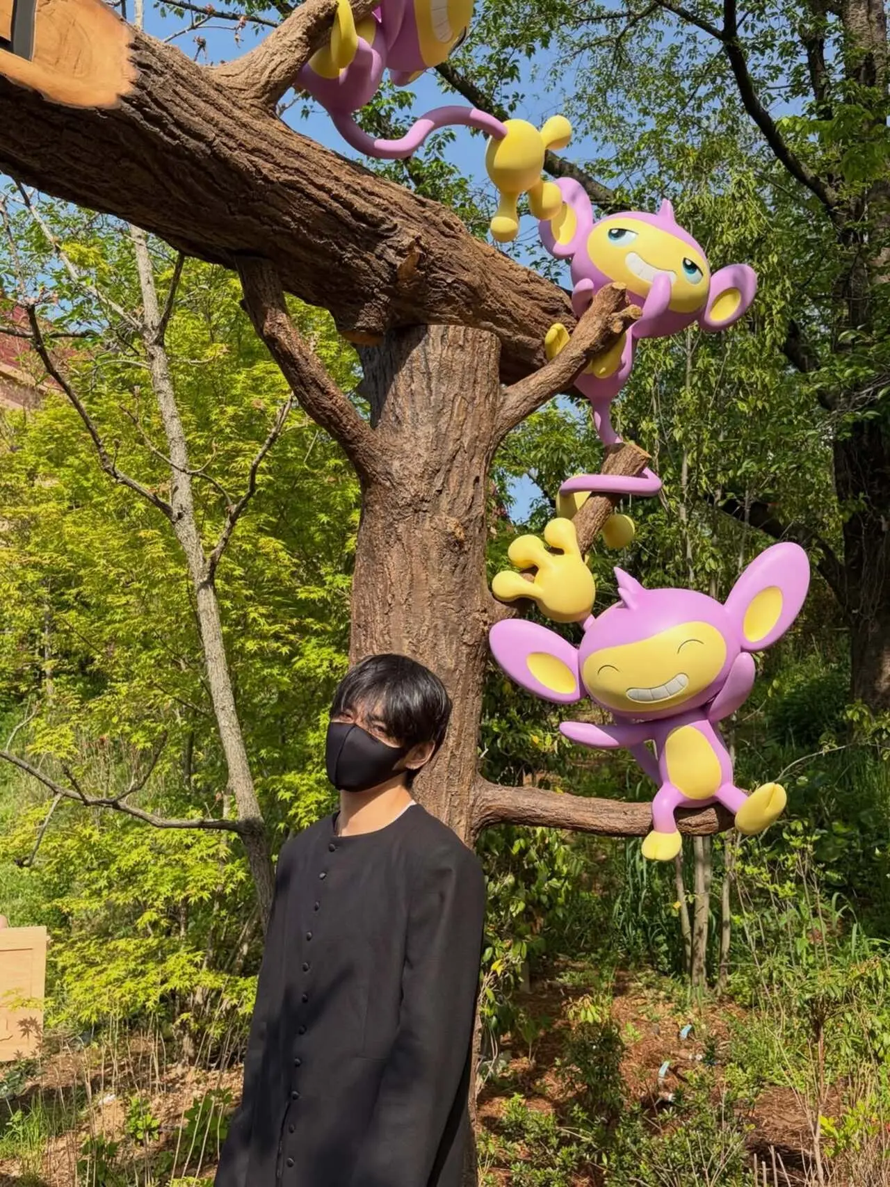 V is captured in a relaxed moment, standing beside playful Pok&eacute;mon figures hanging from a tree. The characters are Aipom&mdash;recognizable by their purple bodies, wide smiles, and distinctive hand-shaped tails.